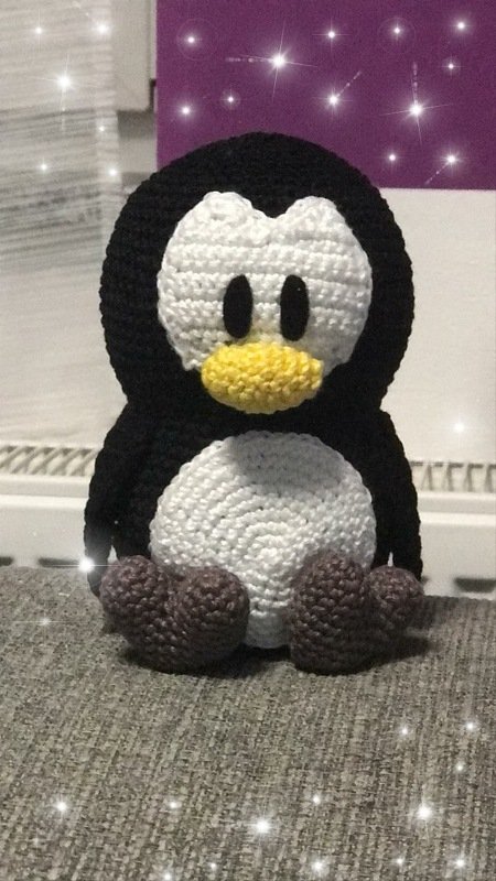 Handgehäkelter Pinguin in Schwarz und Weiß mit gelbem Schnabel und braunen Füßchen auf grauem Stoff
