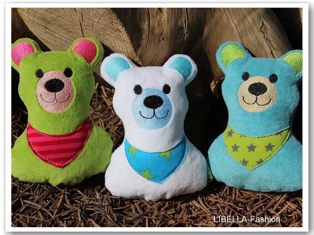 Drei gestickte Kuschelbären in Grün, Weiß und Hellblau mit Halstüchern vor einem Holzstamm auf Nadelholzmulch.