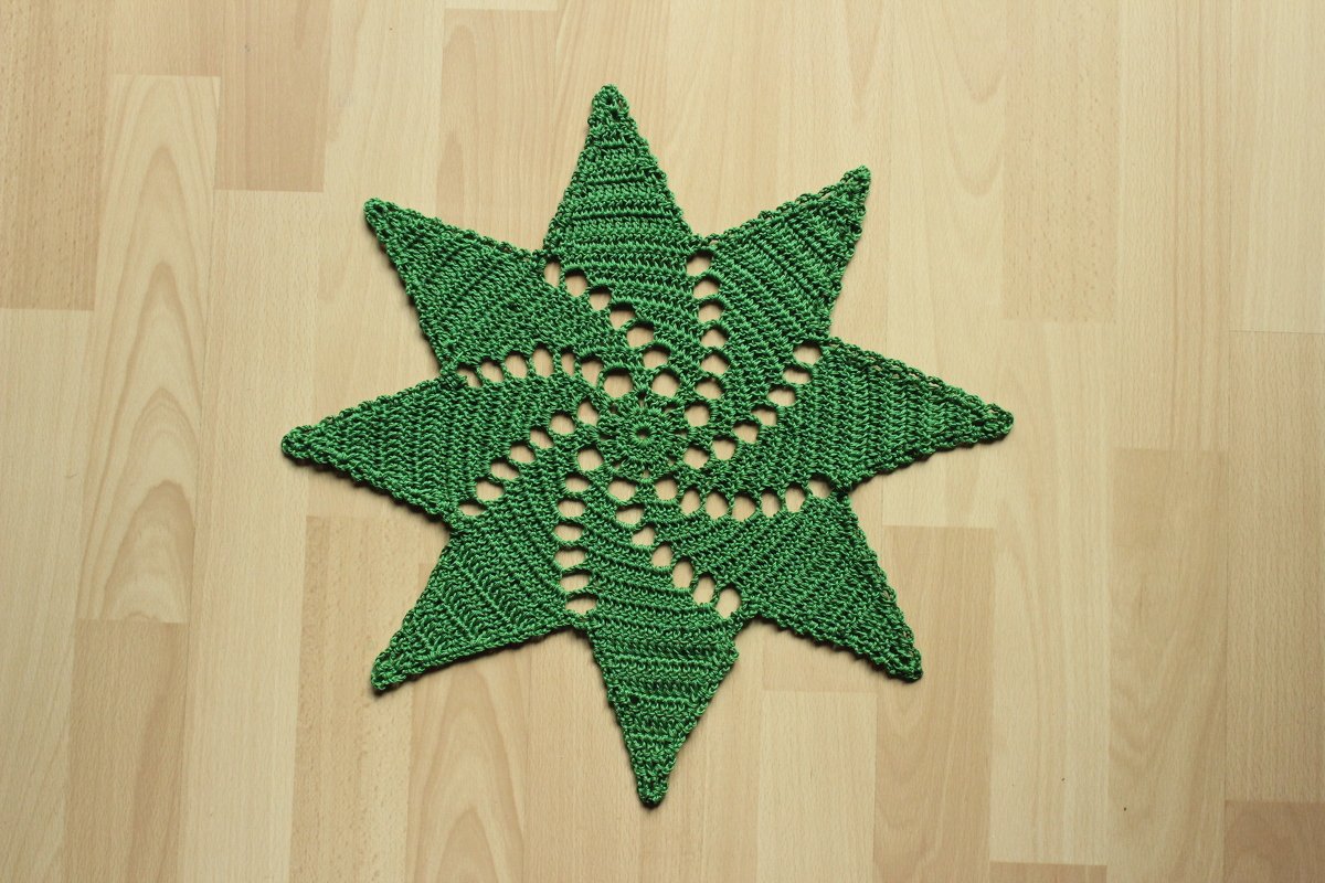 Grünes, gehäkeltes Deckchen in Sternform mit Spiralmuster auf hellem Holzboden