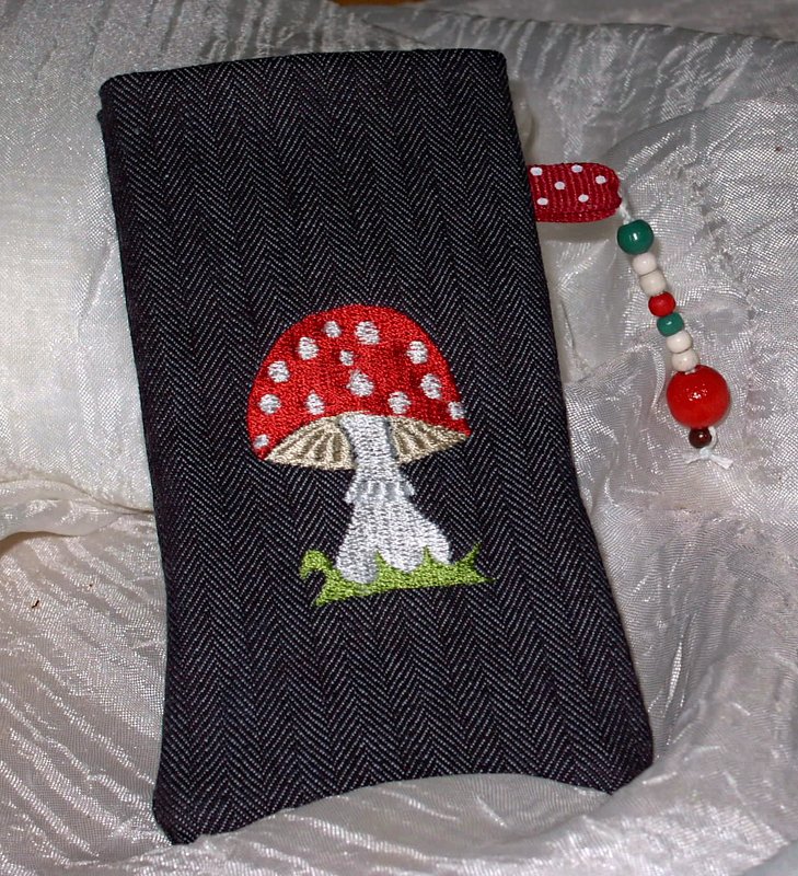 Dunkler Fischgrätstoff mit besticktem roten Fliegenpilz, grünen Grasstichen und Perlenanhänger