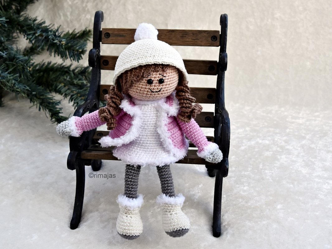 Gehäkelte Amigurumi-Puppe mit braunen Locken, rosa Jacke, weißer Mütze und Stiefeln auf einer Miniaturbank