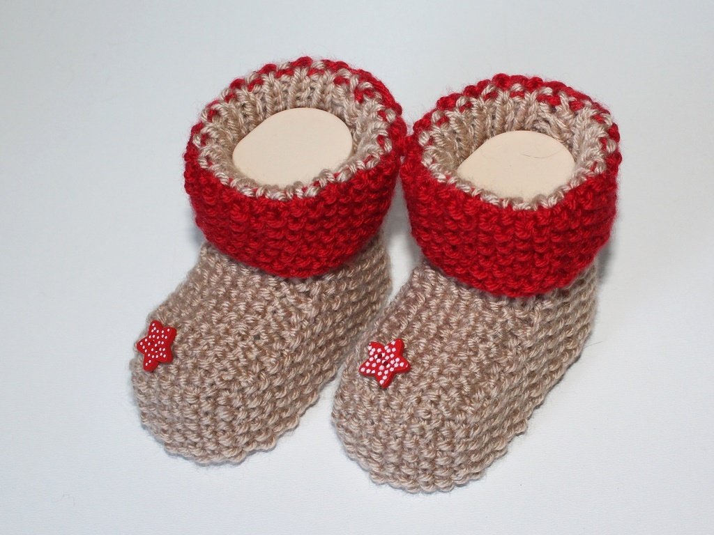 Gestrickte Babyschuhe in Beige mit roten Bündchen und kleinen roten Sternknöpfen auf weißem Hintergrund.
