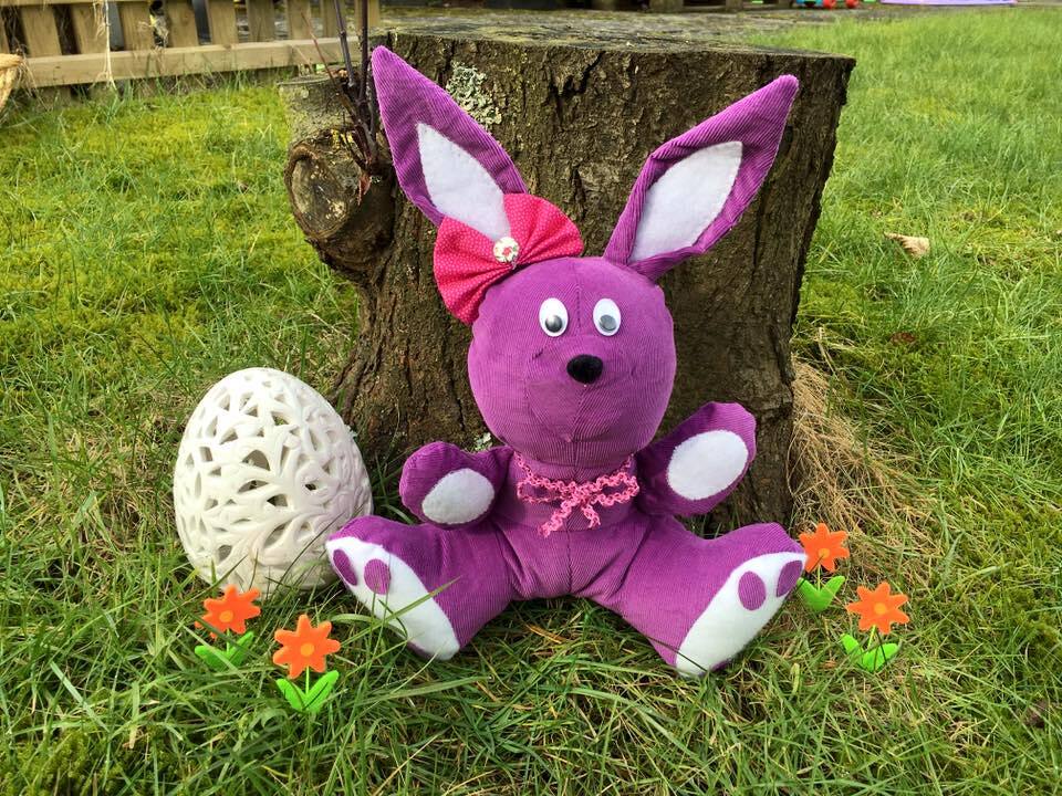 Lila Cord-Stoffhase mit pinker Schleife sitzt im Gras neben einem weißen Deko-Ei.