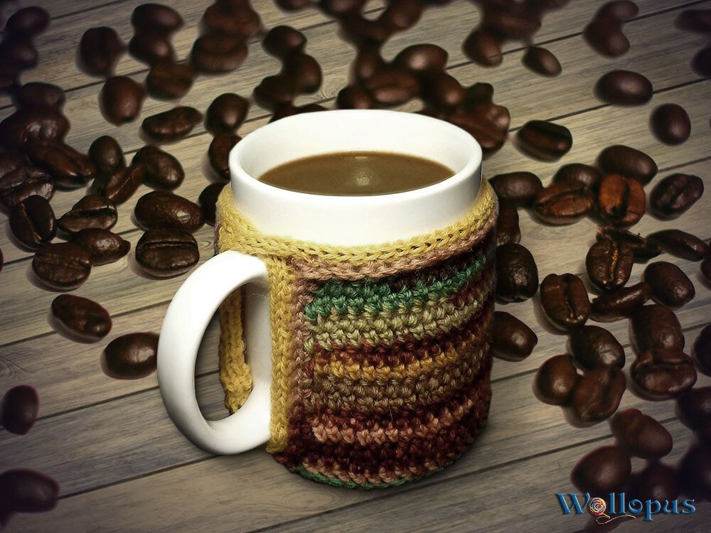 Weiße Kaffeetasse mit braun-grün gestreifter gehäkelter Hülle, umgeben von Kaffeebohnen auf Holztisch.