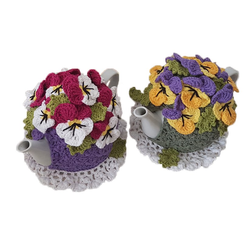 Two handmade crocheted tea cozies with pansy flowers, one purple with pink and white, the other green with yellow and purple, on white doilies.