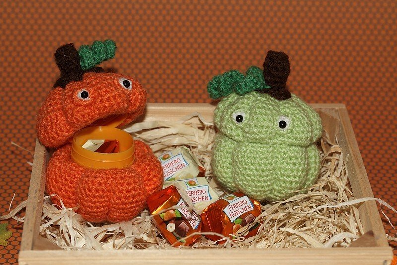 Zwei gehäkelte Kürbisse mit Augen in einer Holzkiste mit Dekostroh und verpackten Pralinen; der orange Kürbis ist geöffnet und zeigt eine gelbe Kunststoffkapsel.