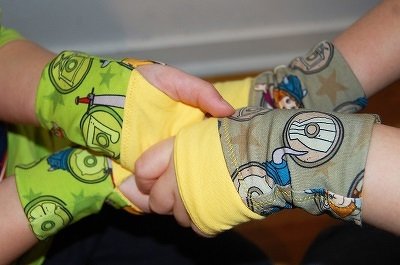 Zwei Kinderhände mit bunten Stoff-Armstulpen mit Comicmotiven und gelben Bündchen, jeweils mit Daumenloch