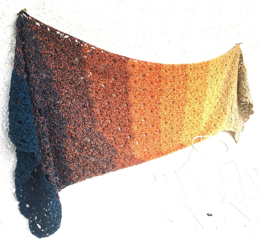 Gehäkelter Schal mit Ombré-Verlauf in Blau, Orange und Beige an einer Wand hängend