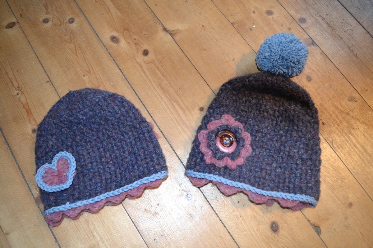 Zwei gehäkelte Damenmützen auf Holzboden, eine mit Herzapplikation, die andere mit Blumenapplikation und Bommel.