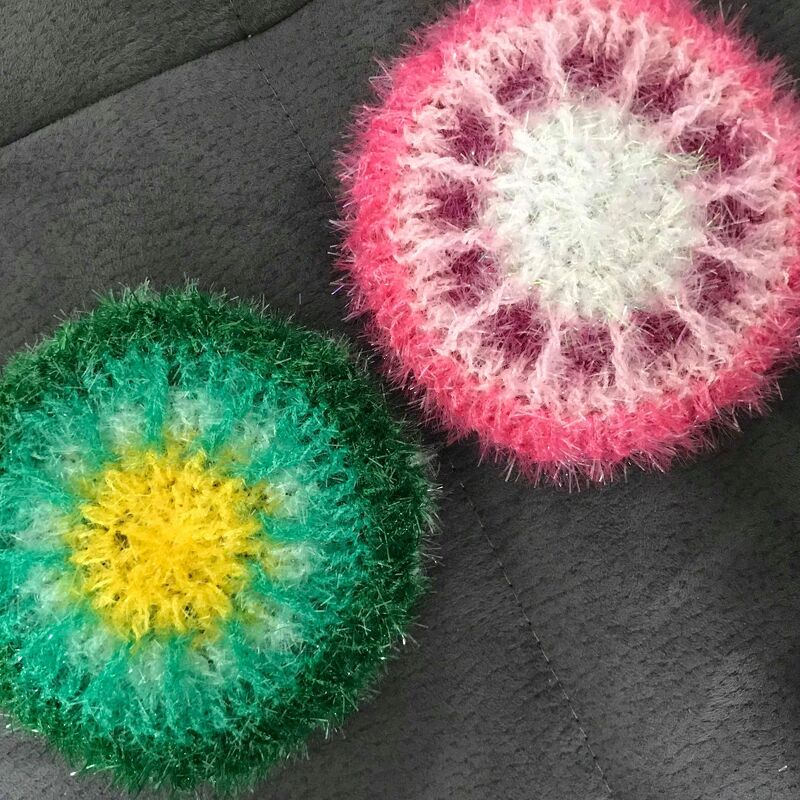 Zwei runde, flauschige gehäkelte Pads in Grün/Gelb und Rosa/Weiß auf dunklem Untergrund