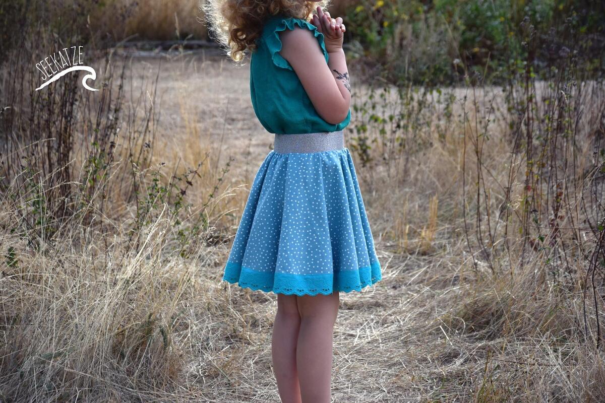 Seitliche Ansicht eines Kindes in hellblauem, weiß gepunktetem Tellerrock mit türkisfarbener Spitze und elastischem Glitzerbund
