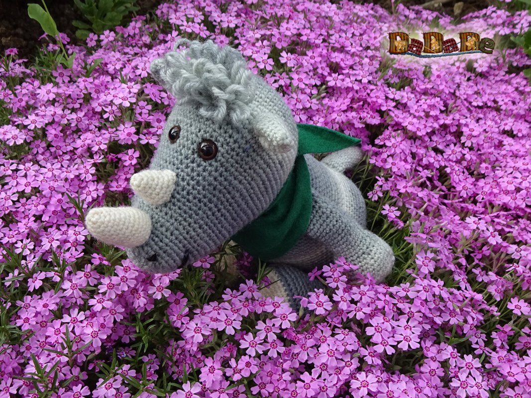 Gehäkeltes graues Nashorn mit weißen Hörnern und grünem Schal auf lila Blumen