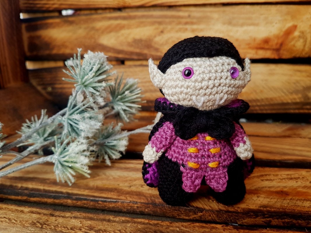 Gehäkelte Amigurumi-Vampirpuppe mit schwarzem Umhang und lila Kleid vor Holzuntergrund