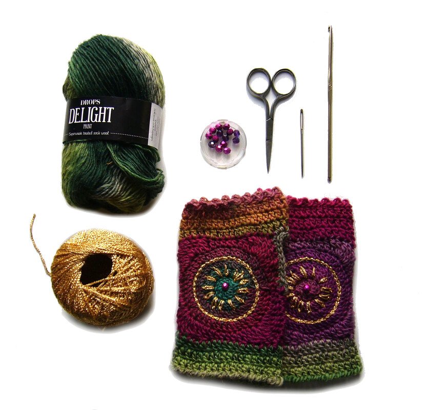 Flat lay of crocheted fingerless gloves with variegated green yarn, metallic gold thread, purple beads, scissors, a crochet hook and needle.