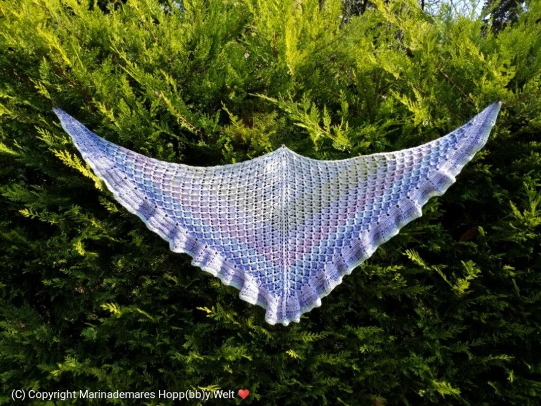 Gehäkeltes Dreieckstuch in Blau-Violett mit Lochmuster und gezackter Bordüre, aufgespannt vor grüner Hecke