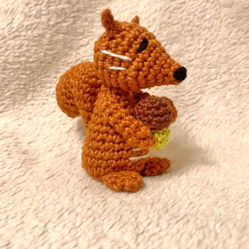 Gehäkeltes braunes Eichhörnchen hält eine Eichel und steht auf hellem, texturiertem Stoff.