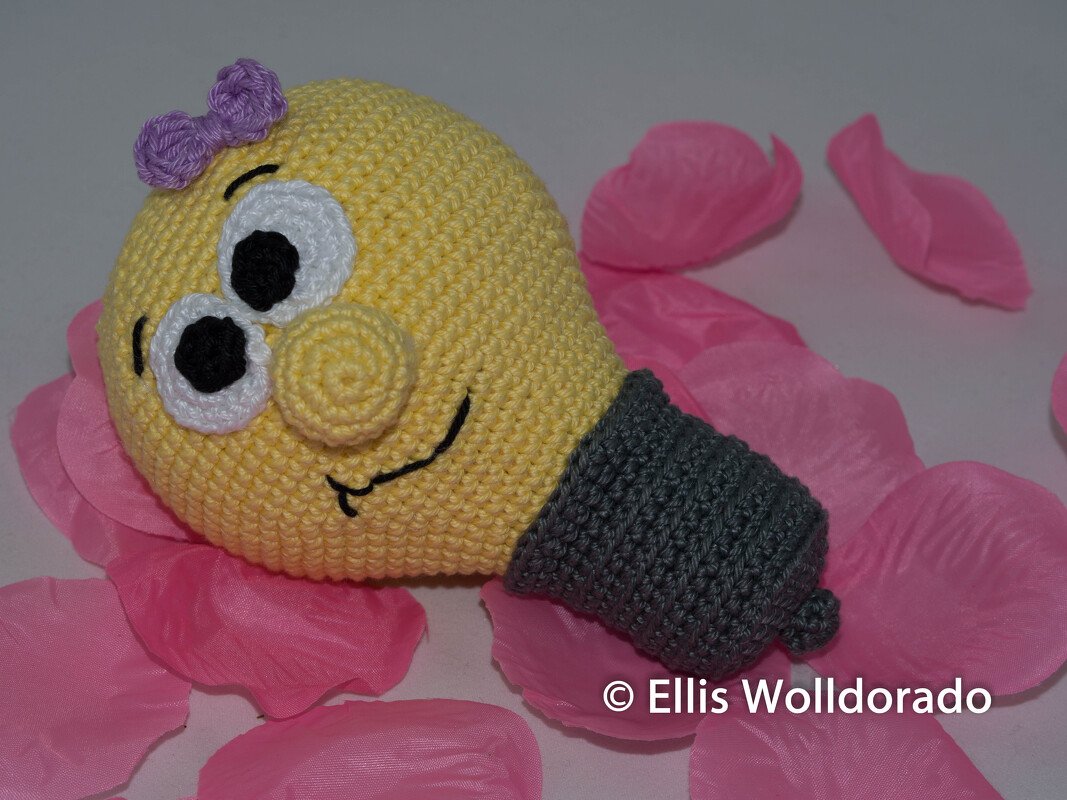 Gehäkelte gelbe Amigurumi-Glühbirne mit grauem Sockel, weißen Augen und lila Schleife auf rosa Blütenblättern
