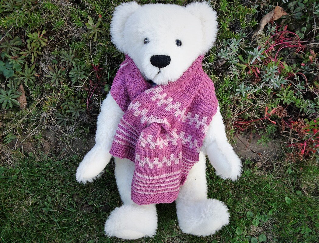 Weißer Plüschteddy mit pink-weiß gemustertem Strickschal und passender Mütze, liegend im Gras.