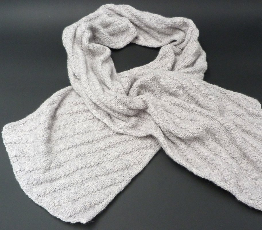Langer hellgrauer gestrickter Schal mit diagonalen Rippen auf dunklem Untergrund