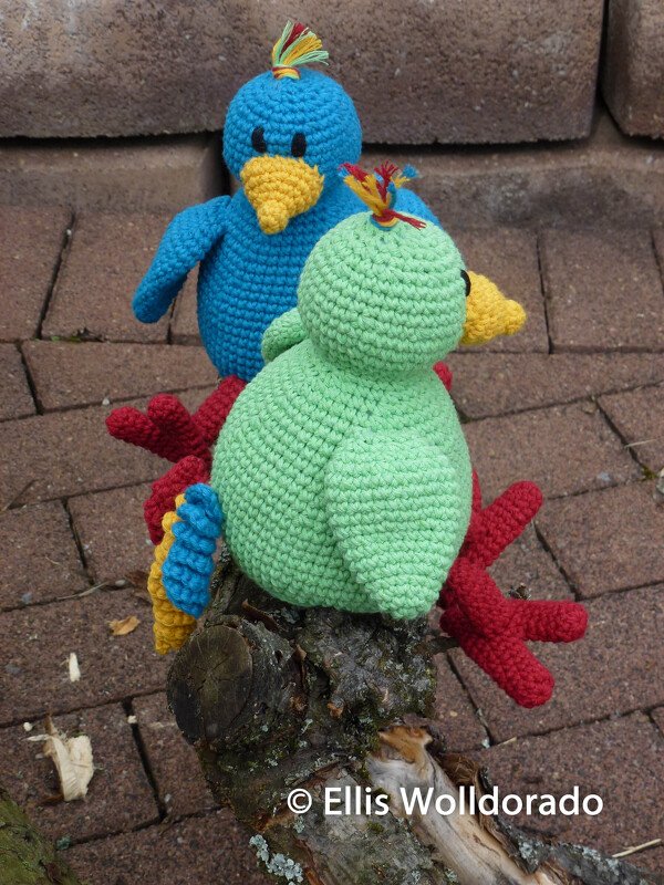 Zwei gehäkelte Amigurumi-Vögel, blau und grün, auf einem Ast vor gepflastertem Untergrund