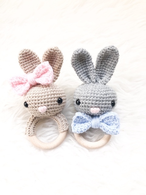 Zwei gehäkelte Hasen-Beißringe mit Holzringen, links beige mit rosa Schleife, rechts grau mit blauer Schleife auf hellem Hintergrund