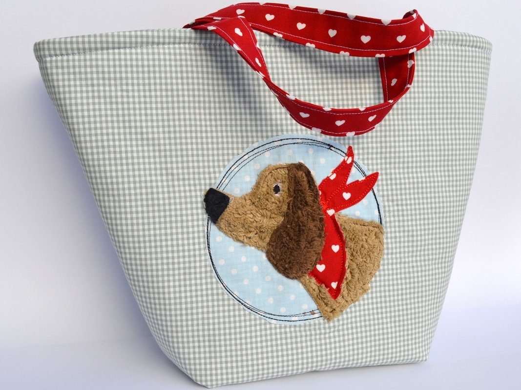 Nahaufnahme einer gestickten Hundeapplikation mit rotem Herz-Halstuch auf hellblauem gepunktetem Kreis vor kariertem Stoff