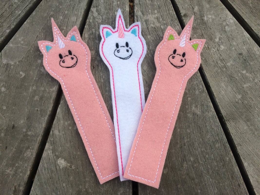 Drei Filz-Einhorn-Lesezeichen in Pastellfarben mit gestickten Konturen auf einem Holztisch