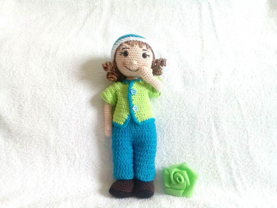 Amigurumi-Puppe mit grünem Cardigan, blauer Hose, braunen Schuhen, lockigem Haar und grüner Häkelrose, stehend auf weißem Untergrund.