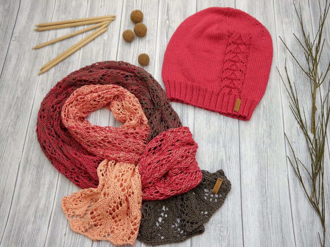 Rote gestrickte Mütze mit Zopfmuster und mehrfarbiger Lochmusterschal auf hellem Holztisch mit Stricknadeln