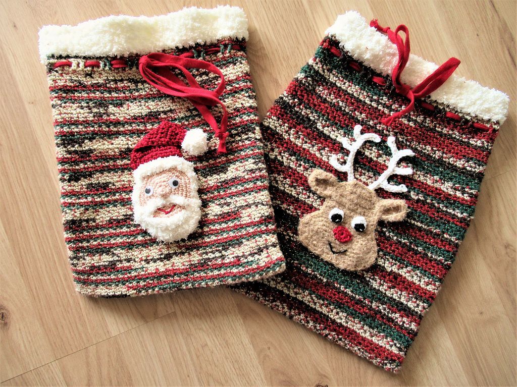 Zwei gehäkelte Nikolaussäckchen mit Weihnachtsmann- und Rentier-Applikationen, gestreiftes Rot‑Grün‑Creme-Muster, Zugband und flauschiger Rand auf Holzboden