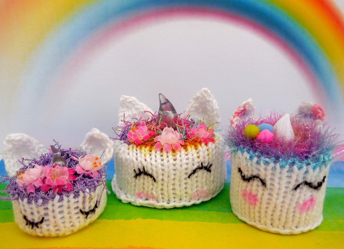 Drei gestrickte Einhörner mit Blumen und Hörnern vor Regenbogenhintergrund