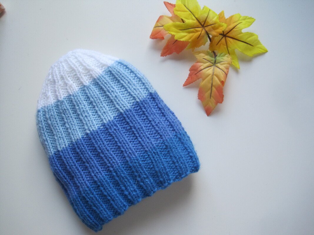 Gestrickte Babymütze in Weiß, Hellblau und Blau liegend auf hellem Untergrund neben künstlichen Herbstblättern