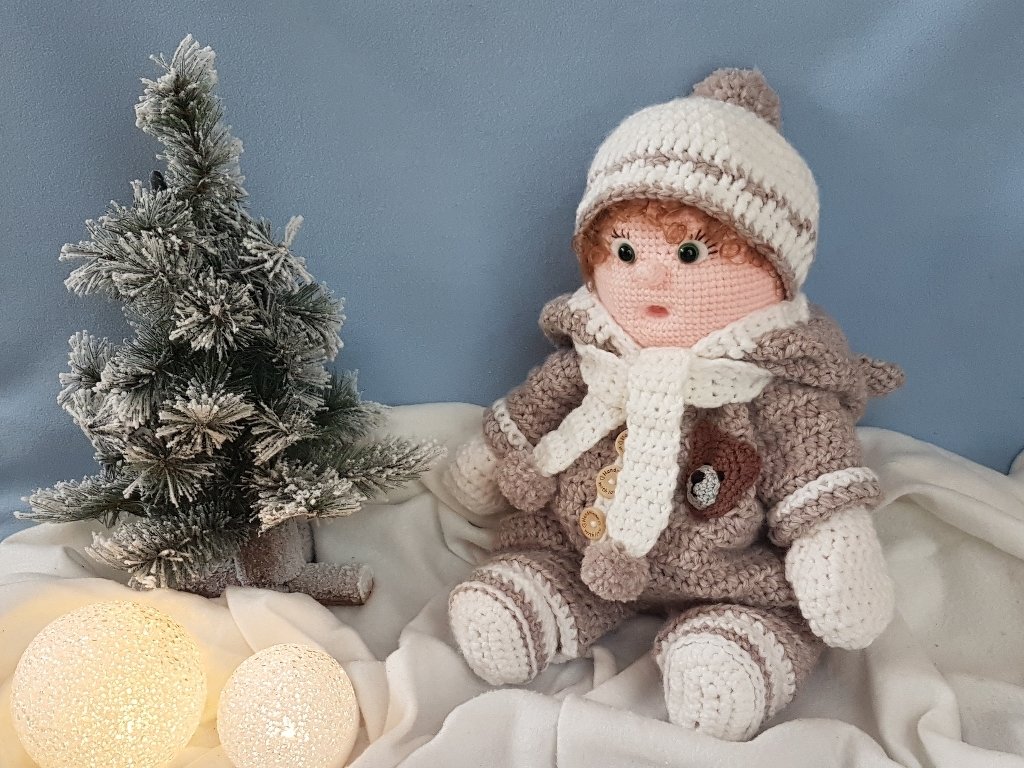 Gehäkelte Amigurumi-Puppe in Winterkleidung mit Mütze und Schal sitzt neben einem kleinen, beschneiten Tannenbaum und leuchtenden Kugeln auf einer weißen Decke