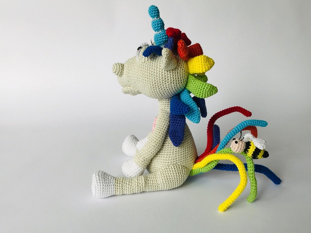 Seitliche Ansicht eines gehäkelten Amigurumi-Einhorns mit Regenbogenmähne und -schweif; am Schweif sitzt eine kleine gehäkelte Biene.