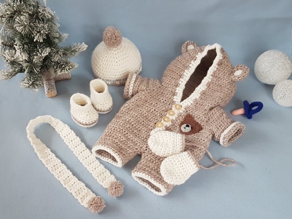 Handgehäkelter Baby-Overall in Beige mit Bärchen-Kapuze, dazu Mütze, Schühchen, Handschuhe und gehäkelte Kordel auf hellem Untergrund.