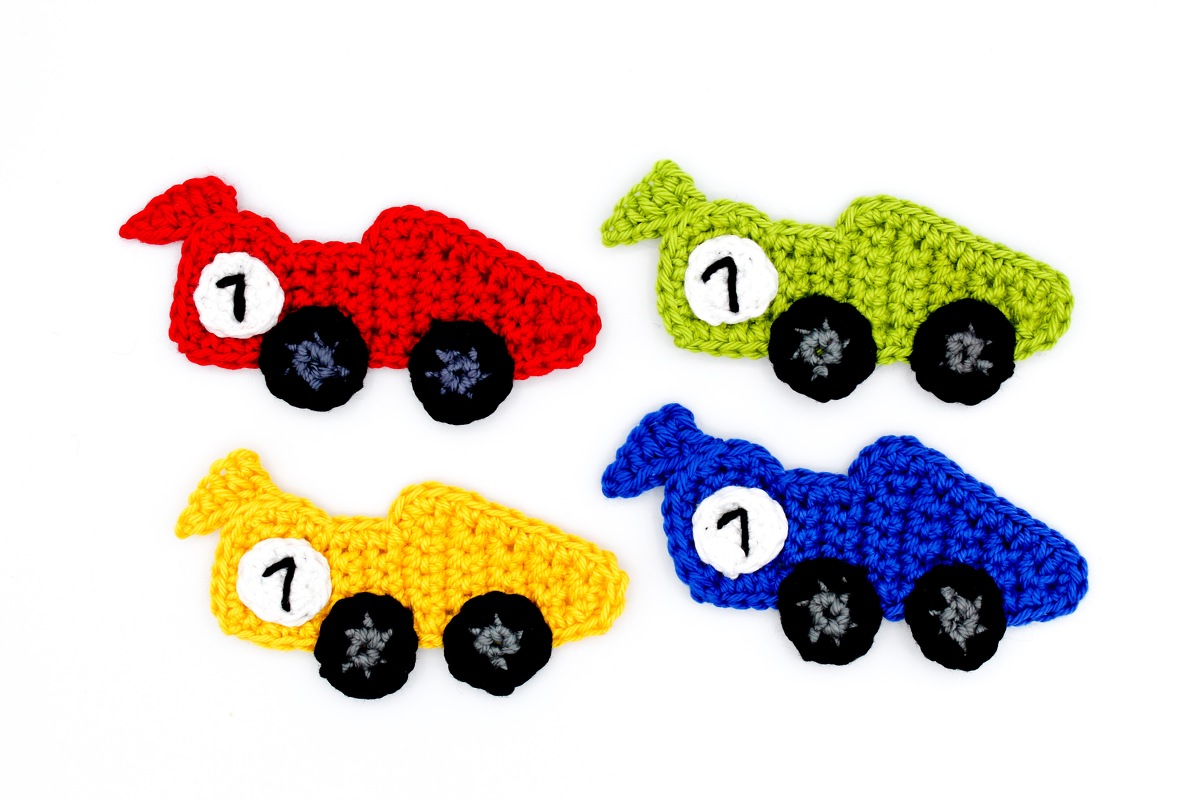 Vier gehäkelte Rennautos in Rot, Grün, Gelb und Blau mit schwarzen Rädern und weißer Startnummer 1 auf weißem Hintergrund.