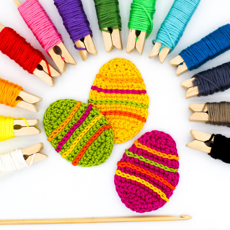 Nahaufnahme: drei gehäkelte Ostereier in leuchtenden Farben, umgeben von auf Holzklammern gewickeltem Garn und einer Häkelnadel.