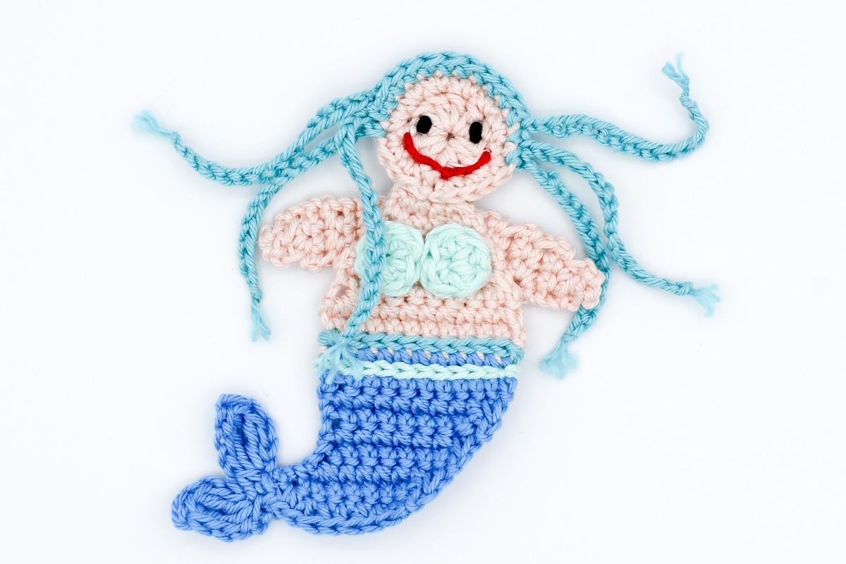 Gehäkelte Meerjungfrau-Applikation mit hellrosa Oberkörper, blauem Schwanz und türkisblauen Zöpfen auf weißem Hintergrund.