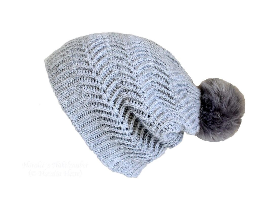 Hellgraue gehäkelte Beanie mit Zickzackmuster und dunklem Bommel auf weißem Hintergrund