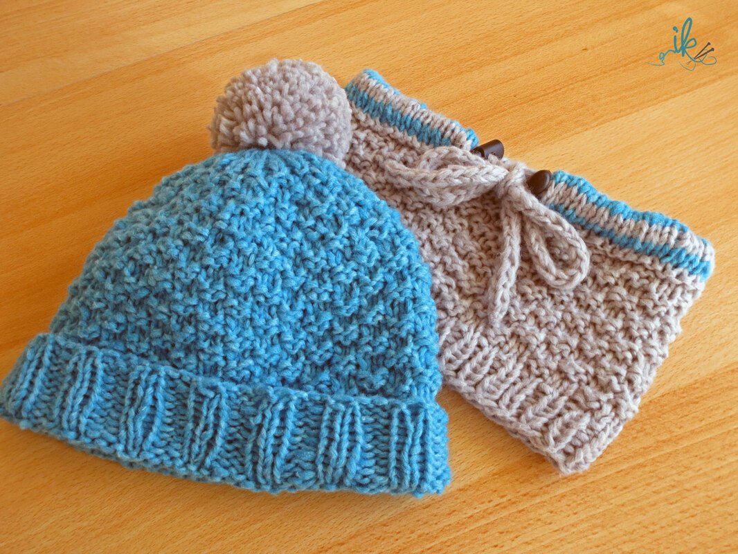 Blaue Kindermütze mit beigem Bommel und beiger Loop‑Schal mit blauer Kante auf Holzuntergrund