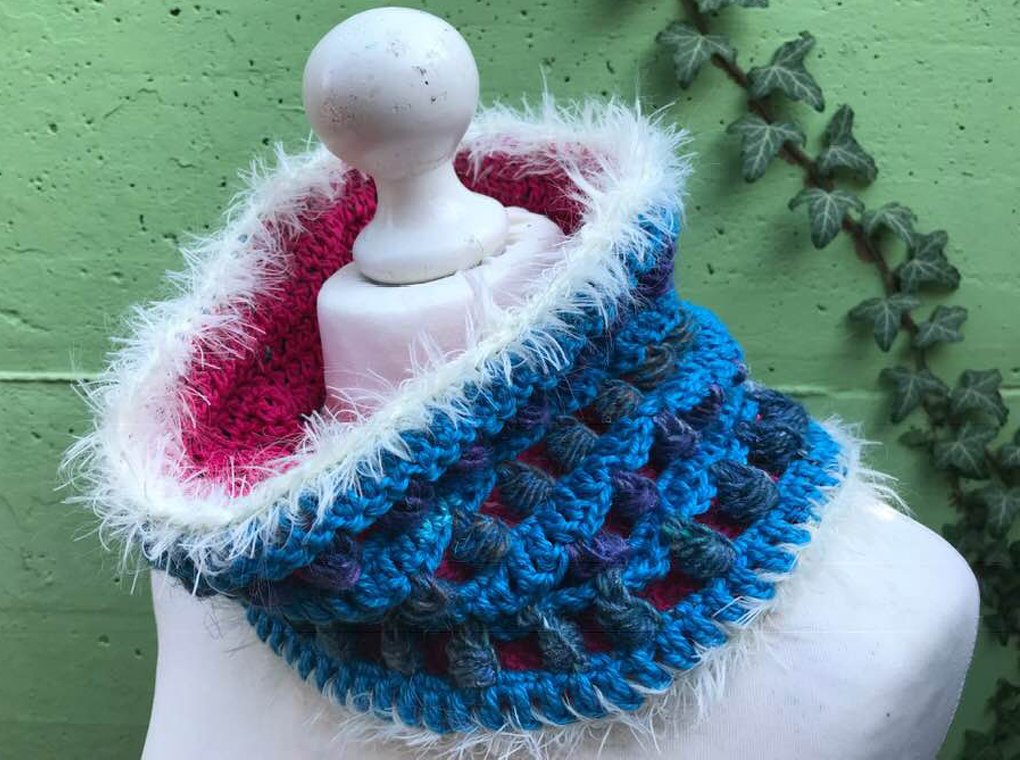 Gehäkelter Loop in blau-türkis mit pinker Innenlage und weißem Plüschrand auf Schneiderbüste vor grüner Wand