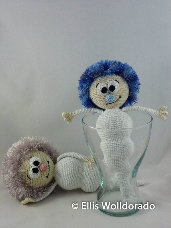 Zwei gehäkelte Amigurumi-Babys mit flauschigem Haar, eines in einem Glas, das andere liegend vor hellem Hintergrund
