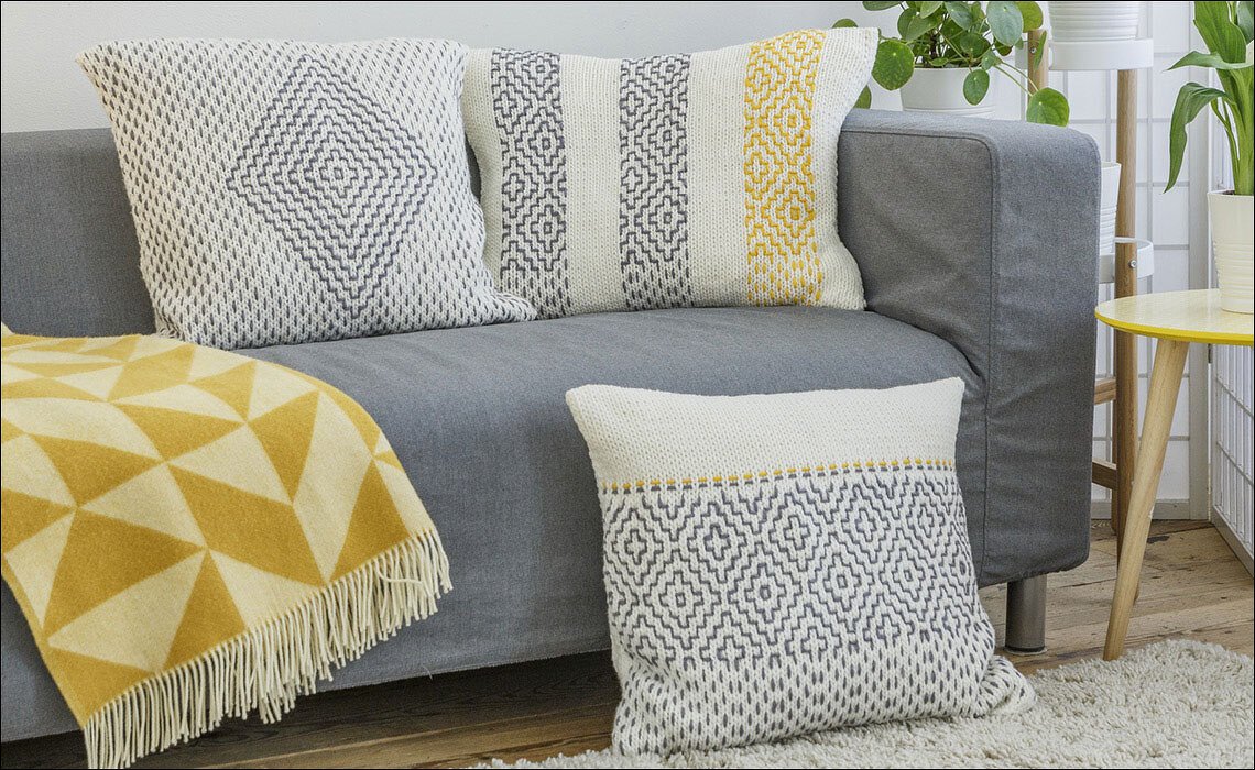 Drei gestrickte Kissen und eine Decke auf einem grauen Sofa in Weiß, Grau und Gelb mit geometrischen Mustern.