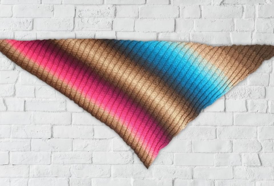 Striped gradient crochet triangle shawl in pink, brown, and blue laid flat on a white brick wall.
