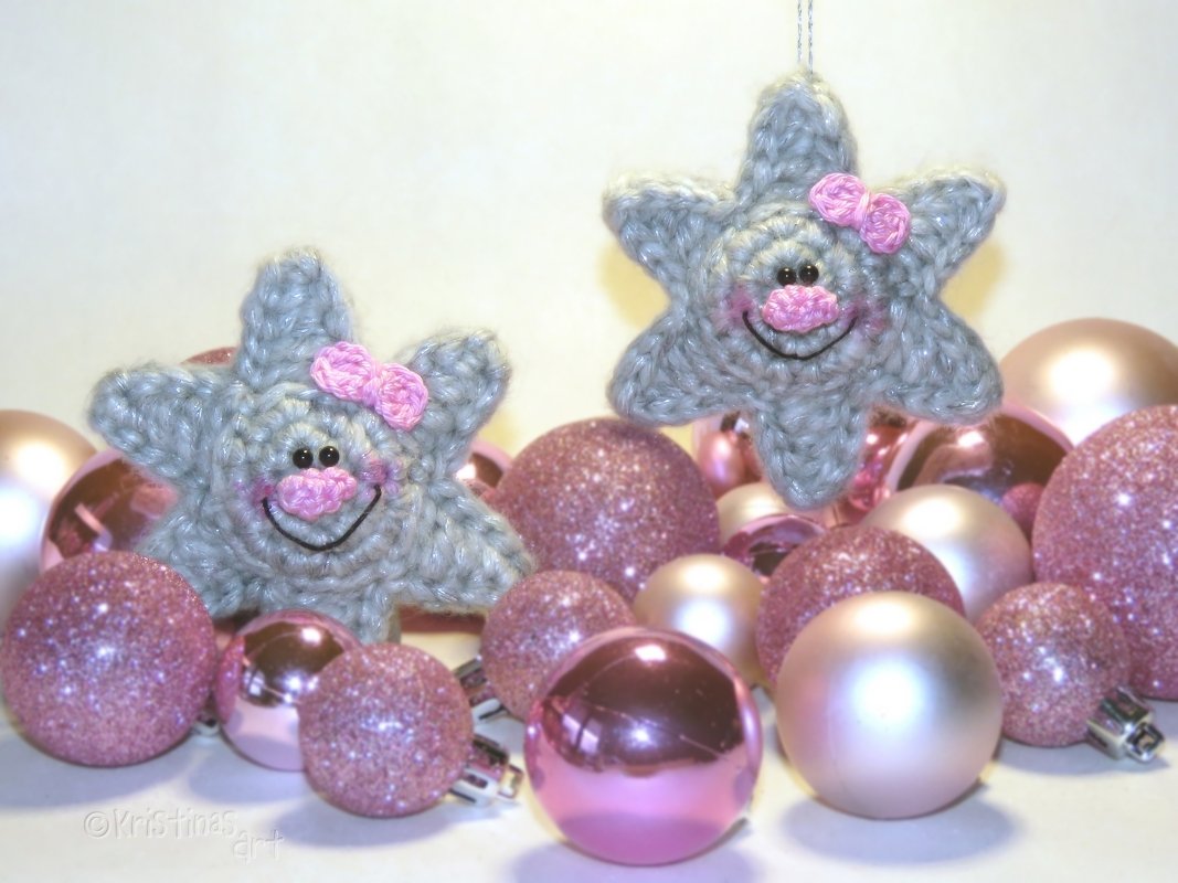 Zwei gehäkelte graue Sterne mit rosa Schleifen und lächelnden Gesichtern, umgeben von rosa Weihnachtskugeln.