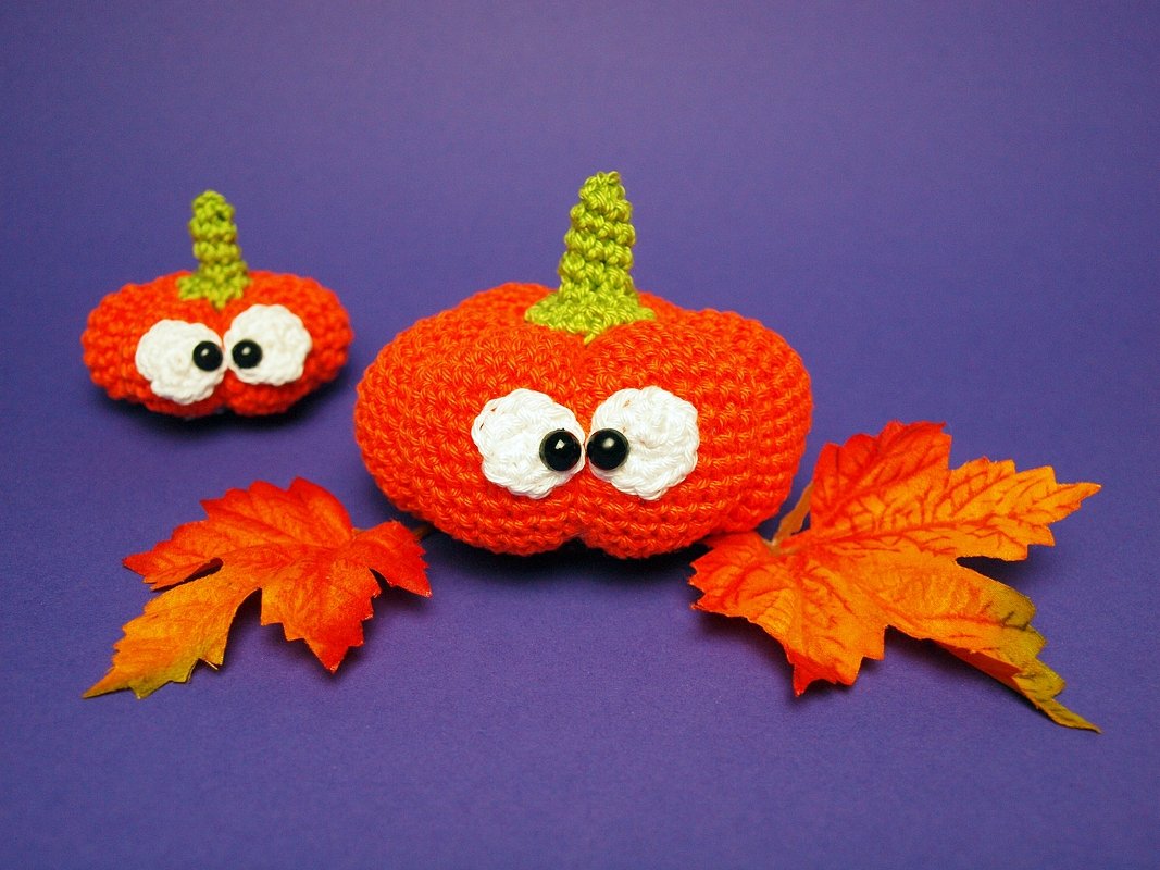 Zwei orange gehäkelte Kürbisse mit Wackelaugen und grünen Stielen, umgeben von Herbstblättern auf violettem Hintergrund.