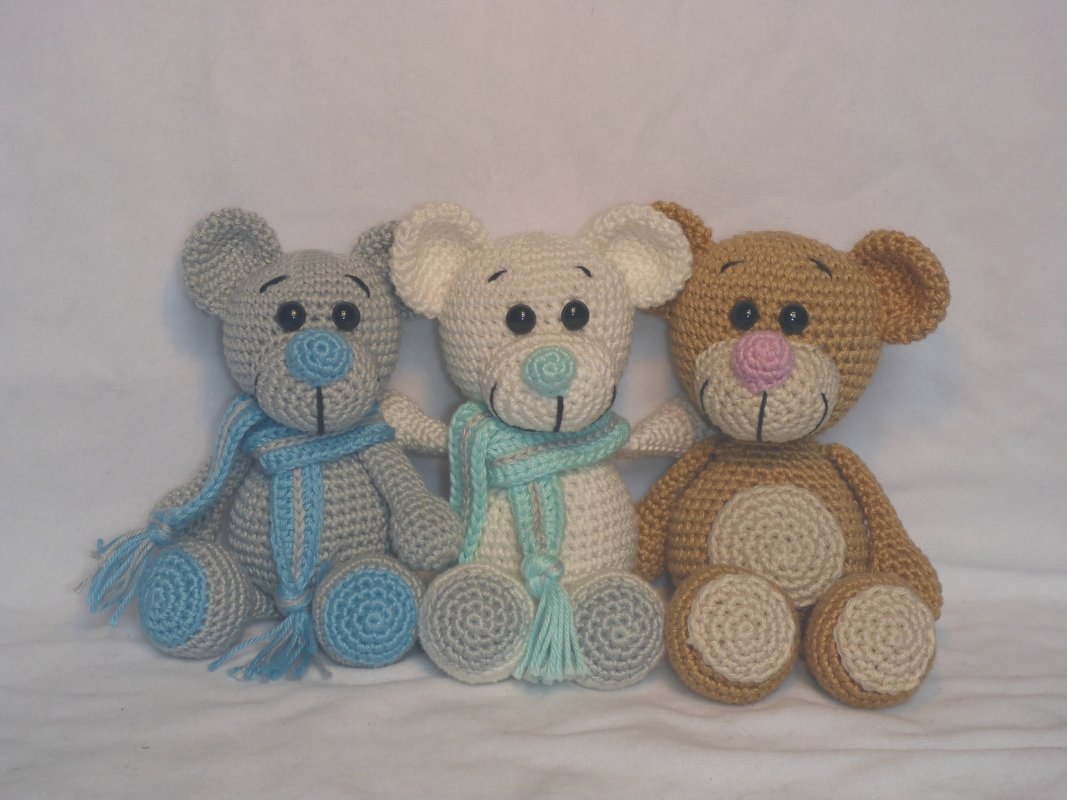 Drei gehäkelte Teddybären sitzend: grau mit blauem Schal, creme mit türkisfarbenem Schal, hellbraun mit rosa Nase.