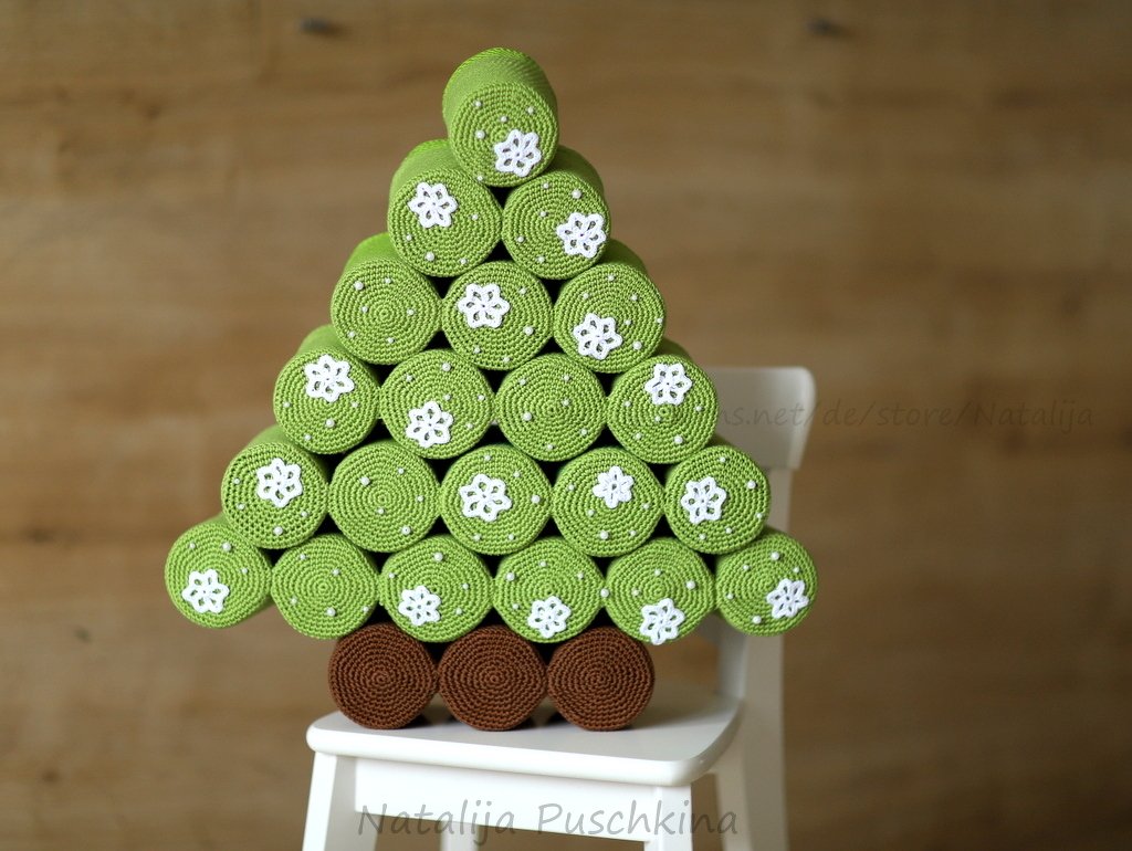 Gehäkelter Tannenbaum aus grünen Rollen mit weißen Blumenapplikationen und brauner Basis auf einem weißen Stuhl vor Holzwand