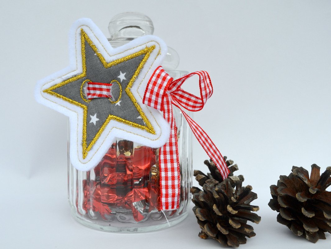 Gestickter Stern-Anhänger aus Filz mit goldener Umrandung und rotkariertem Band an einem Glas mit roten Bonbons, daneben Tannenzapfen