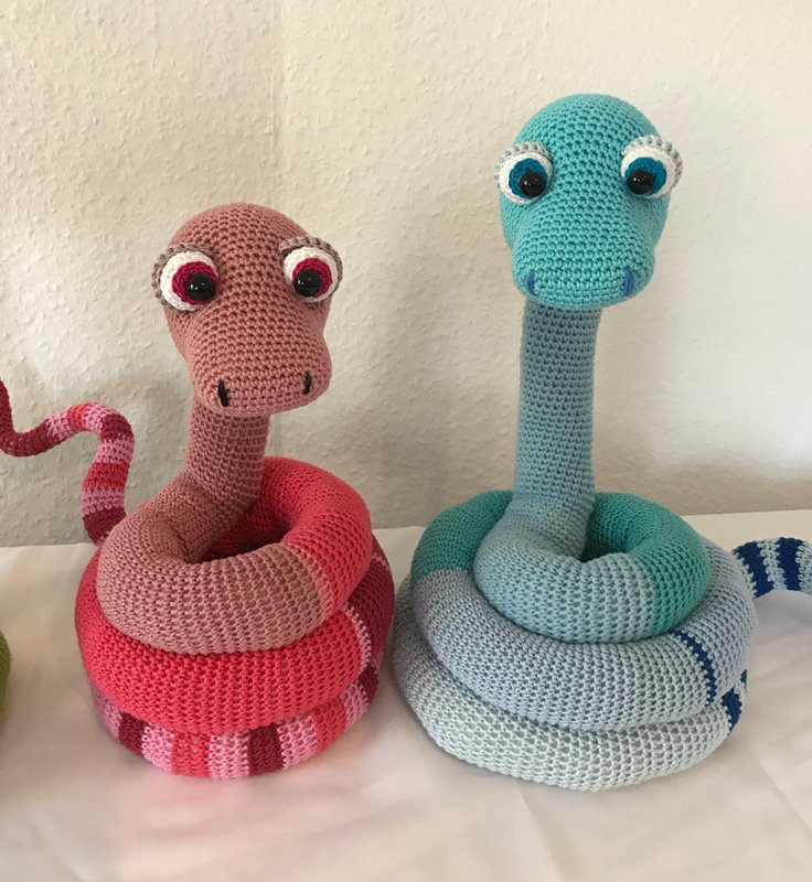 Zwei aufgerollte gehäkelte Amigurumi‑Schlangen, rosa und blau, mit großen Augen und gestreiftem Körper.